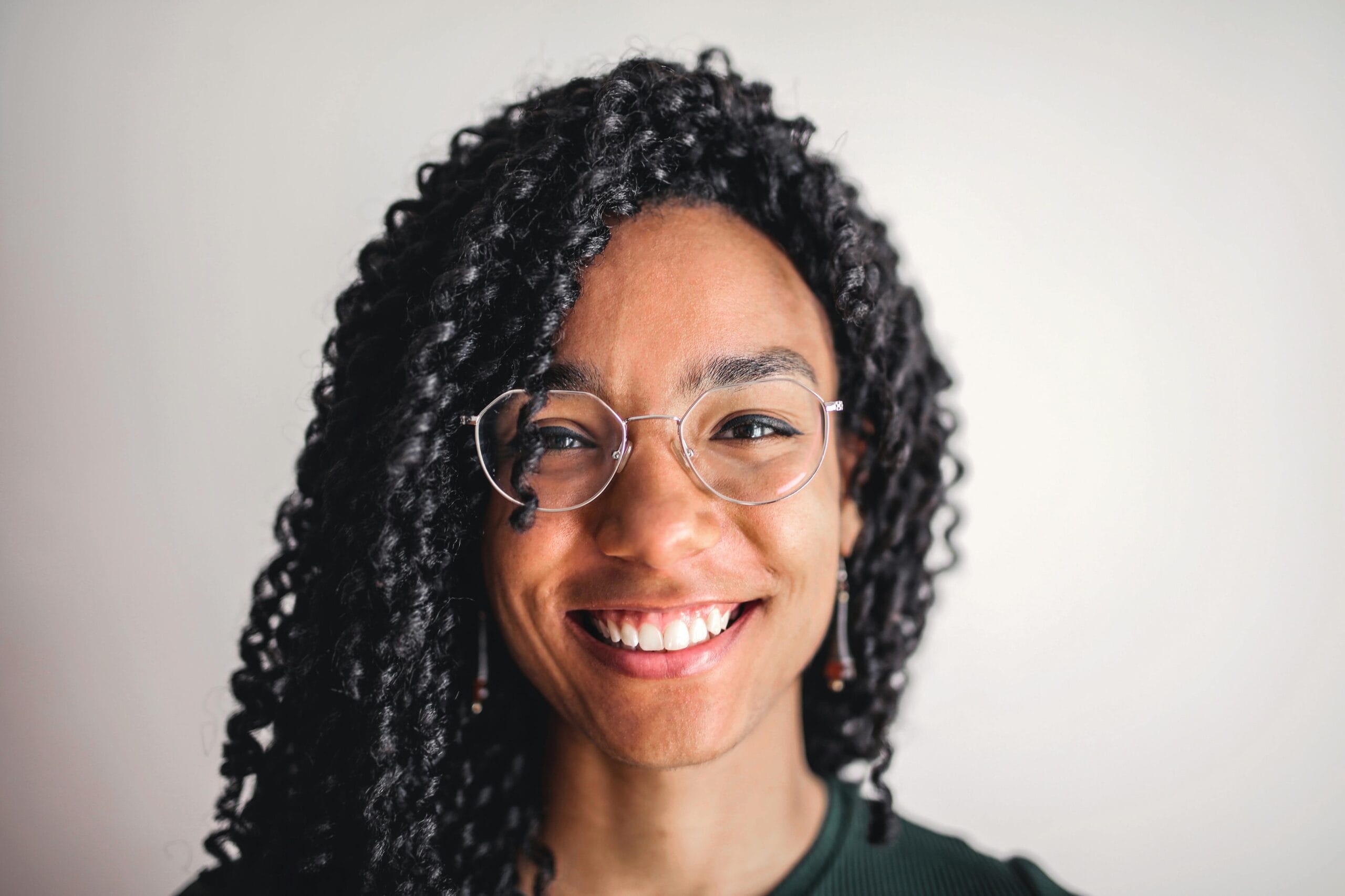 Close-up portrait of a young woman with curly hair and glasses smiling indoors.